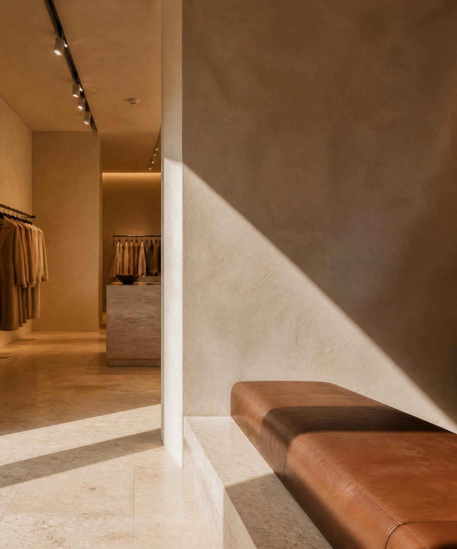 Interior de loja premium com luz natural diagonal sobre parede de reboco e banco de couro caramelo